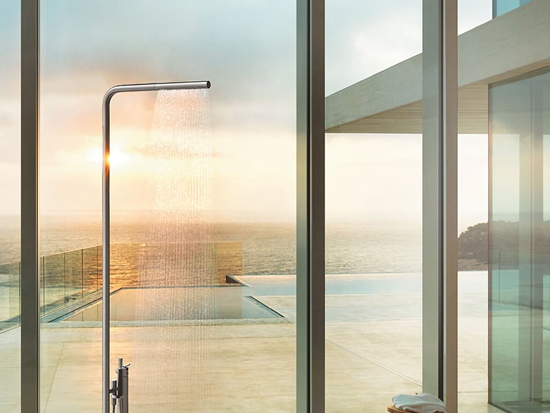A VOLA shower head against a window with a sunset behind it